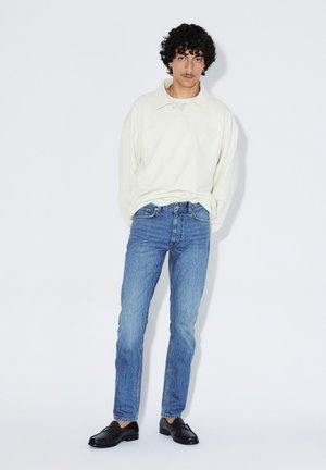 Junger Mann, der vor weißem Hintergrund steht, trägt ein weißes Poloshirt mit langen Ärmeln, blaue Jeans und schwarze Loafers, die Hände in den Taschen, blickt nach vorne.