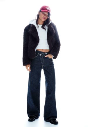 Mladá žena v tmavej kožušinovej bunde cez bielu krátku mikinu s kapucňou, široké tmavé rifle, čierne topánky a červená šiltovka, pózujúca s jednou rukou v vrecku.