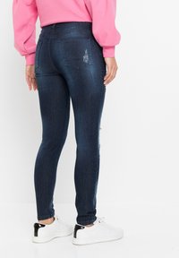 Tmavě modré skinny džíny s hladkou denimovou texturou, které mají dvě zadní kapsy a mírné odřené detaily, doplněné bílými teniskami.