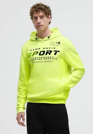 Mann mit lockigem Haar, der eine hellgelb-grüne Kapuzenjacke mit schwarzem Text und schwarze Hose trägt, steht vor einem einfarbigen hellen Hintergrund.