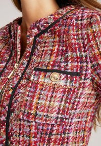 Geweven tweed jacket in rood, goud en meerdere kleuren. Voorzien van een rits aan de voorkant, zwarte accenten en een decoratieve pocket met details in goudkleurig metaal.
