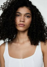 Modella con capelli neri ricci, che indossa un top bianco con texture, a spalline larghe e un dettaglio con bottoni davanti.