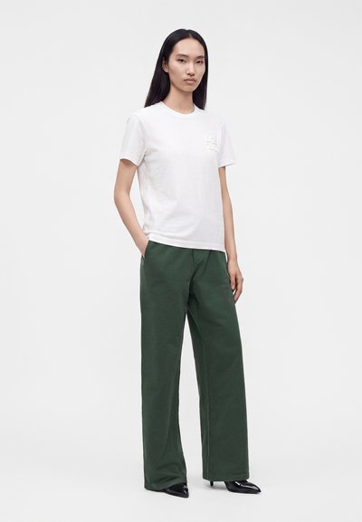 Femme debout aux longs cheveux foncés portant un t-shirt blanc à manches courtes, un pantalon large vert foncé et des chaussures noires pointues.
