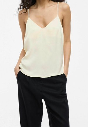 Femme portant un haut crème sans manches à col en V avec fines bretelles et pantalon noir, mains dans les poches, sur un fond uni.