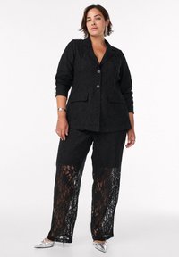 Mujer vistiendo un blazer de encaje negro y pantalones de encaje transparente a juego, combinados con flats plateados de punta y pendientes llamativos.