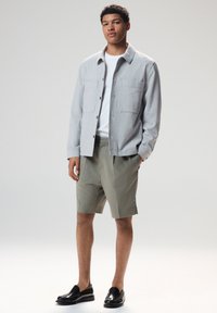 Gray textured jacket with two chest pockets, white T-shirt, olive shorts, and black loafers. Model stands in a neutral background.