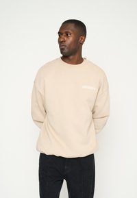Beiger Sweatshirt mit oversized Passform, Rundhalsausschnitt und gesticktem Logo auf der Brust. Kombiniert mit schwarzen strukturierten Hosen.