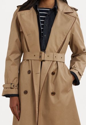 Beiger doppeltreihiger Trenchcoat mit Gürtel, geknöpfte Manschetten, getragen über einem gestreiften Hemd und einem dunklen Oberteil mit Knöpfen.