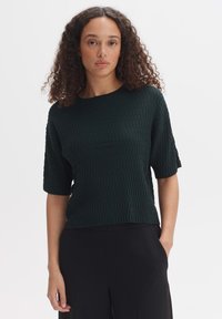 Haut court à manches courtes, côtelé et vert foncé, avec un col rond, présentant une texture structurée et une coupe décontractée. Assorti à un pantalon noir.