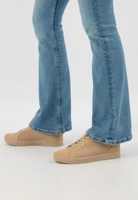 Sneakers in tela beige con superficie strutturata, occhielli dorati e suole in gomma, abbinati a jeans a zampa d'elefante di un blu chiaro.