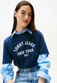 Camiseta de algodón azul marino con estampado blanco "TOMMY JEANS NEW YORK CITY", superpuesta sobre una camisa de cuello azul claro. Se lleva con pantalones cortos de mezclilla de cintura alta.