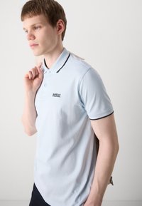 Polo din bumbac de culoare albastru deschis, cu guler cu borduri bleumarin și un logo pe față. Textură fină, mâneci scurte și design ajustat.