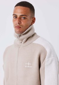 Hombre con pelo corto que lleva un suéter de cremallera beige y blanco con cuello alto y el logo de PXF en el pecho, contra un fondo blanco liso.