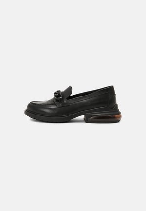 Zwart leren loafer met grove zool, decoratieve ketting op de wreef en transparante luchtgeveerde hak, zijaanzicht op witte achtergrond.