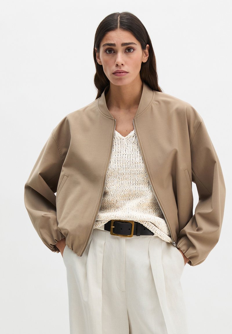 Woman wearing a tan bomber jacket over a cream knit sweater with white pleated trousers and a black belt with gold buckle.