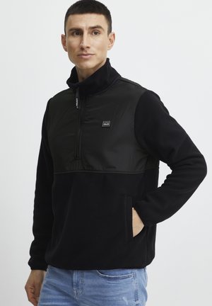 Black fleece pullover with a zippered collar and a smooth nylon panel at the chest. Includes a front pocket and a small logo patch.