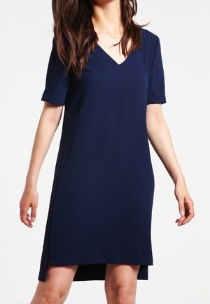 Femme portant une robe bleu marine mi-genoux à manches courtes et col en V, debout avec les bras détendus le long du corps.