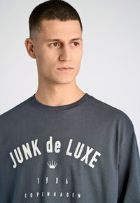 Camiseta de manga corta gris oscuro con el texto blanco "JUNK de LUXE 1986 COPENHAGUE" y un gráfico de una corona, hecha de una mezcla de algodón.