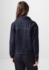 Giacca in denim scuro con maniche lunghe e colletto. Presenta cuciture a contrasto e una silhouette leggermente accorciata. Visuale posteriore mostrata.