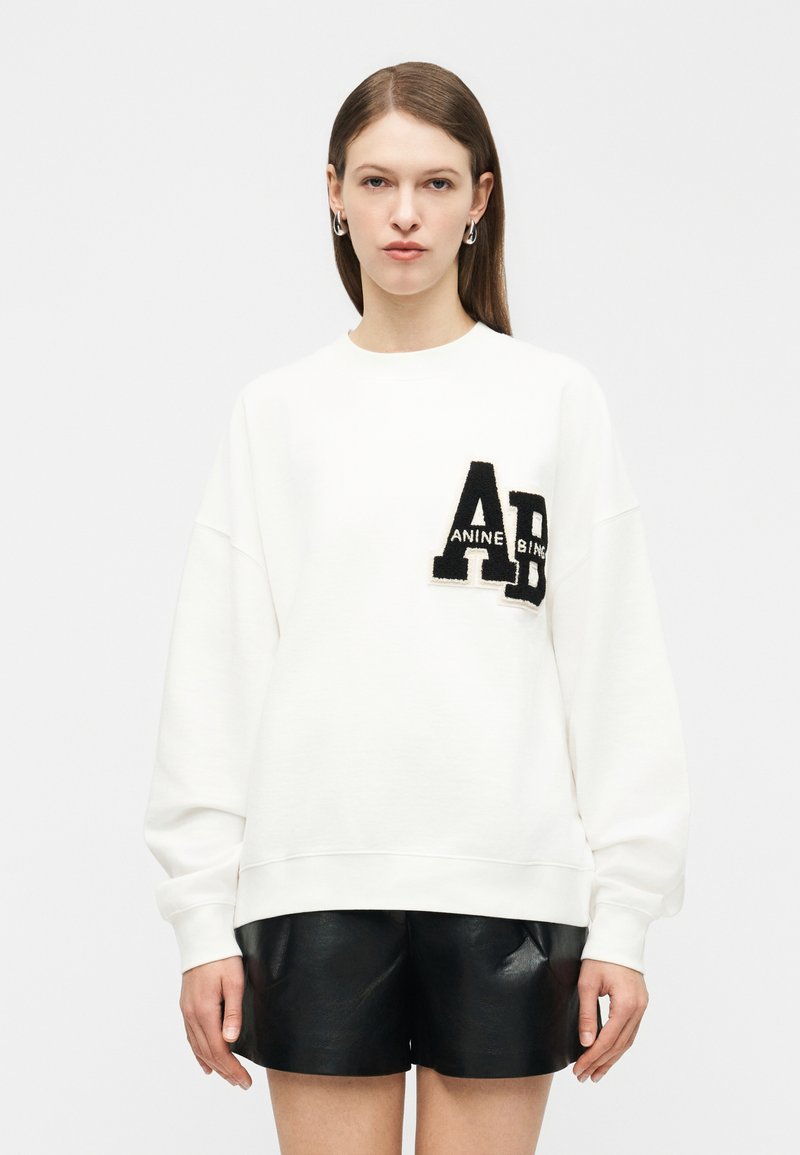 Γυναίκα με ίσια καστανά μαλλιά φορά λευκό φούτερ με μαύρό τσίπ "AB" και μαύρο δερμάτινο σορτς, στέκεται μπροστά σε απλό φόντο.