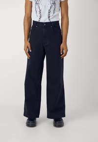 Široké tmavě modré džínové kalhoty s pěti kapsami, zapínáním na knoflík a viditelnými švy, v kombinaci se světlým tričkem s potiskem.