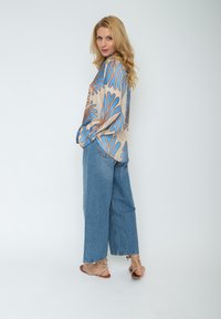 Beige Bluse mit langen Ärmeln und blauen sowie orangefarbenen abstrakten Mustern, kombiniert mit hellblauen weiten Jeans und flachen Sandalen.