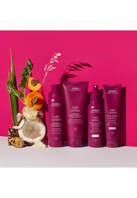 Produits de soin capillaire Aveda Color Control présentés sur un fond rose vif. Les contenants sont violets avec des étiquettes décrivant l'utilisation du produit.