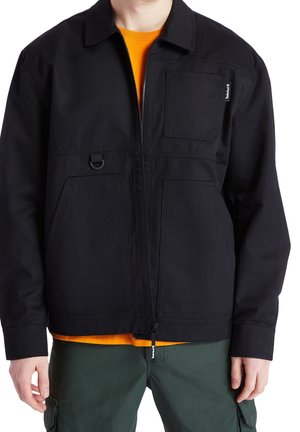 Sort jakke lavet af holdbart stof med krave, frontlynlås, flere lommer og et lille logo-tags. Båret over en orange skjorte.