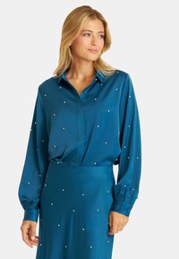 Blusa de satén color verde azulado con mangas largas, cierre de botones en la parte delantera y adornos similares a perlas en toda su superficie. Textura suave, diseño fluido.