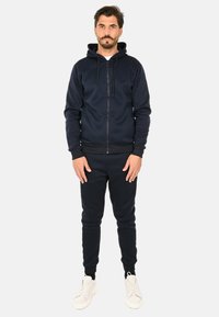 Felpa con zip color blu navy e coulisse, pantaloni in felpa a gamba tapered coordinati e sneaker bianche. Il tessuto è liscio e elasticizzato.