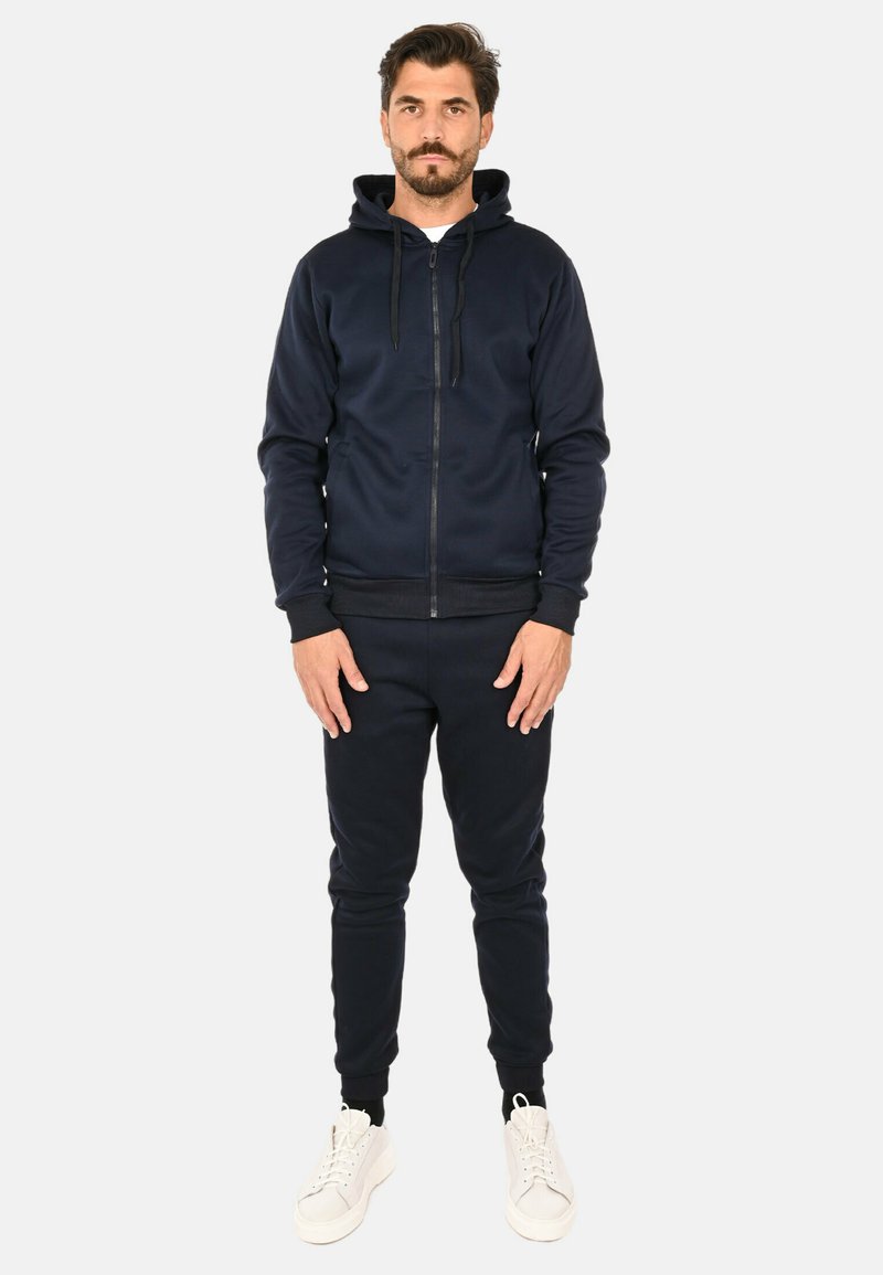 Felpa con zip color blu navy e coulisse, pantaloni in felpa a gamba tapered coordinati e sneaker bianche. Il tessuto è liscio e elasticizzato.