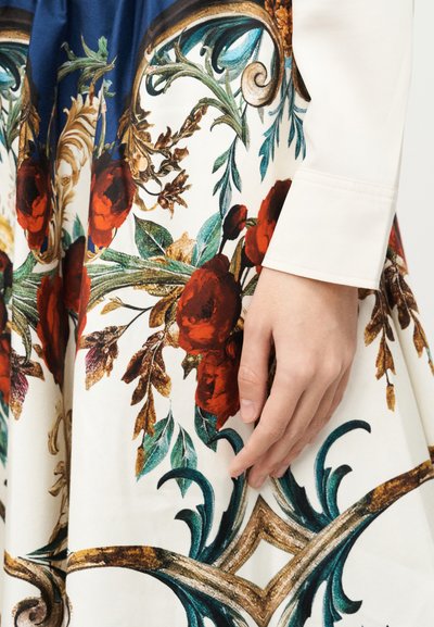 Main reposant sur une robe ornée de motifs floraux et de volutes complexes en rouge, vert, bleu et or sur un tissu blanc et bleu.