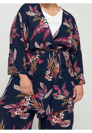 Persoon die een marineblauwe, bloemen kimono-stijl jas en bijpassende broek draagt, vastgebonden in de taille, over een wit topje, afgebeeld van nek tot midden van het dijbeen.