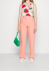 Vrouw draagt een mouwloze trui met aardbeienpatronen, koraalroze broek, pastelkleurige sneakers en houdt een kleine groene handtas vast.