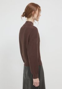 Frau mit roten Haaren in einem tiefen Dutt, die einen braunen Pullover mit langen Ärmeln und einen schwarzen Rock trägt, seitlich vor einem hellen, einfarbigen Hintergrund.