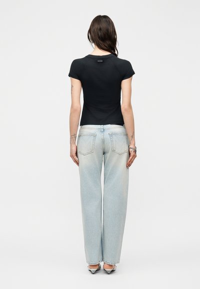 Γυναίκα με την πλάτη γυρισμένη φοράει μαύρο εφαρμοστό κοντομάνικο τοπ, τζιν παντελόνι με φαρδιά πόδια σε ανοιχτή απόχρωση και παπούτσια με τακούνι σε απλό φόντο.