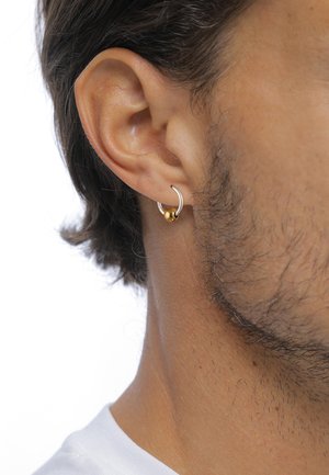 Boucle d'oreille en argent avec une perle dorée, portée sur le lobe. Le teint de la peau est visible, avec des cheveux mi-longs et une barbe courte.