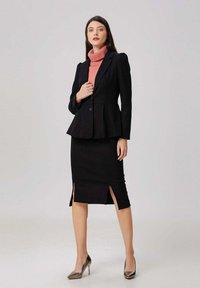 Femme portant un blazer noir sur un col roulé rose, une jupe noire longueur genoux avec des fentes à l'avant, et des escarpins métaliques à bout pointu, se tenant debout.