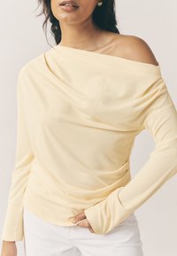 Haut à manches longues drapé jaune avec un design épaules nues, une texture douce et des manches ajustées, associé à un pantalon blanc.