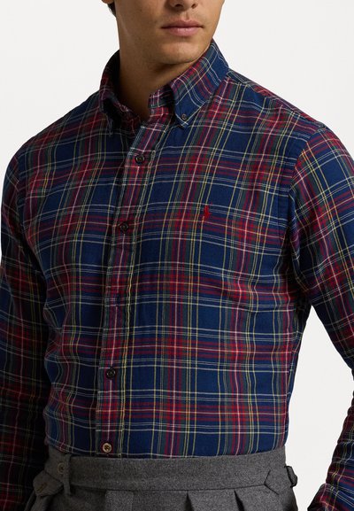 Chemise en coton à carreaux rouge, marine et jaune avec un col boutonné et un petit logo sur la poitrine gauche. Présente des boutons foncés.