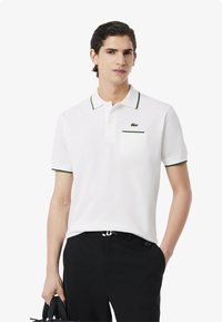 Polo bianca con accenti verdi, maniche corte, colletto con bottoni e taschino sul petto con logo. Indossata con pantaloni neri.