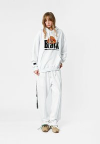 Sudadera blanca con la palabra "Black" y un gráfico de un oso, combinada con pantalones jogger blancos con cremalleras. El modelo lleva zapatillas de tonos neutros.