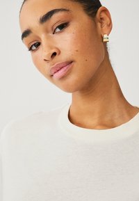 T-shirt blanc à col rond, texture lisse, et gros plan sur le visage d'une personne portant des boucles d'oreilles en or et un maquillage naturel.