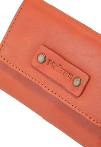 Orangenes Lederportemonnaie mit strukturierter Oberfläche, ausgestattet mit einem Metall-Druckknopfverschluss und geprägtem Etikett "MAÎTRE". Genahtes Kanten sind sichtbar.