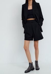 Completo nero su misura composto da blazer e pantaloncini, abbinato a un top corto nero e stivaletti grigi lucidi con lacci e dettagli testurizzati.