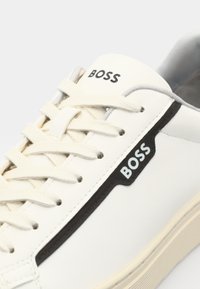 Baskets blanches avec des accents noirs. Fabriquées en cuir lisse, elles présentent un bout arrondi, des lacets texturés et un logo "BOSS" bien visible.