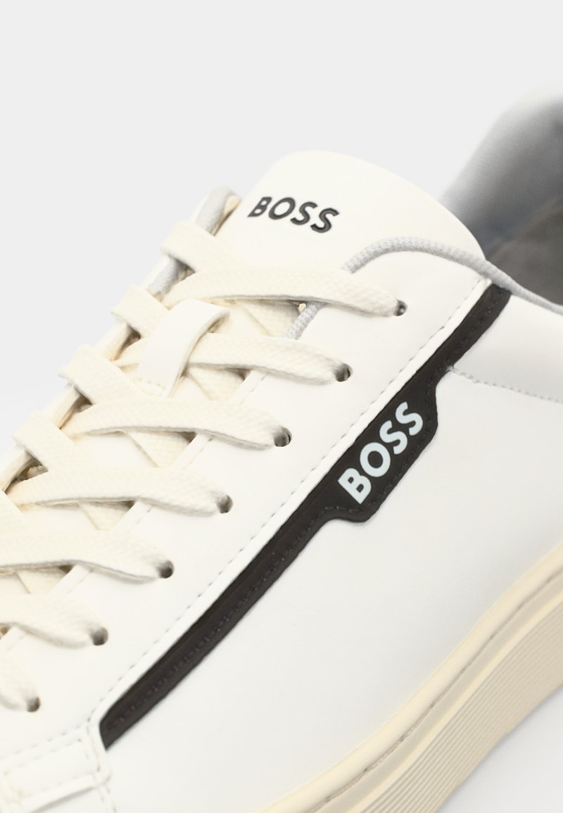 Baskets blanches avec des accents noirs. Fabriquées en cuir lisse, elles présentent un bout arrondi, des lacets texturés et un logo "BOSS" bien visible.