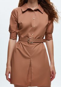 Robe chemise marron avec des manches bouffantes courtes, fermeture à boutons, taille ceinturée avec une boucle circulaire et fente à l'avant pour plus de détails.