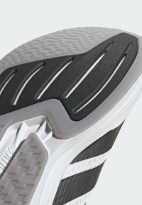 Semelle de chaussure en caoutchouc noir texturé avec des accents blancs et des rainures en motif, se transitionnant vers un bord blanc lisse.