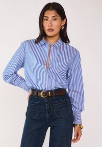 Camicia a righe blu e bianche con colletto, caratterizzata da maniche lunghe e una vestibilità rilassata, indossata con jeans scuri a vita alta e una cintura marrone.
